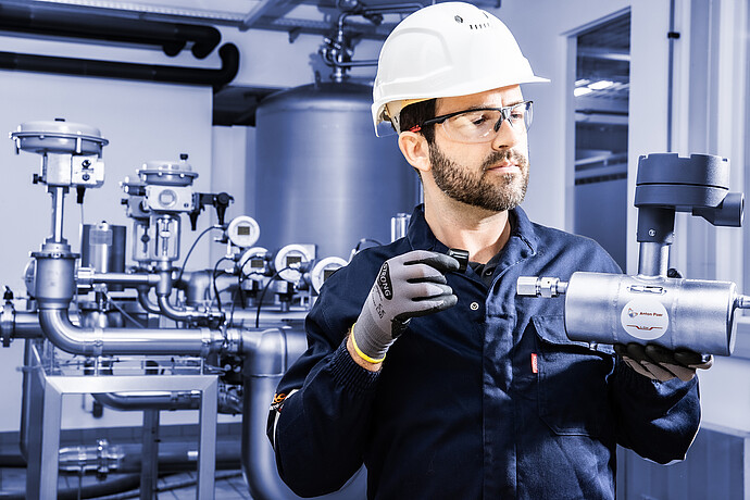 Coriolis flow meter L-Cor 8000 held by a technician in protective gear, standing in an industrial piping system with valves and gauges.