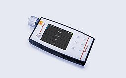 Anton Paar Cora 101 handheld Raman spectrometer lying on a flat surface with its display showing scan menu options.