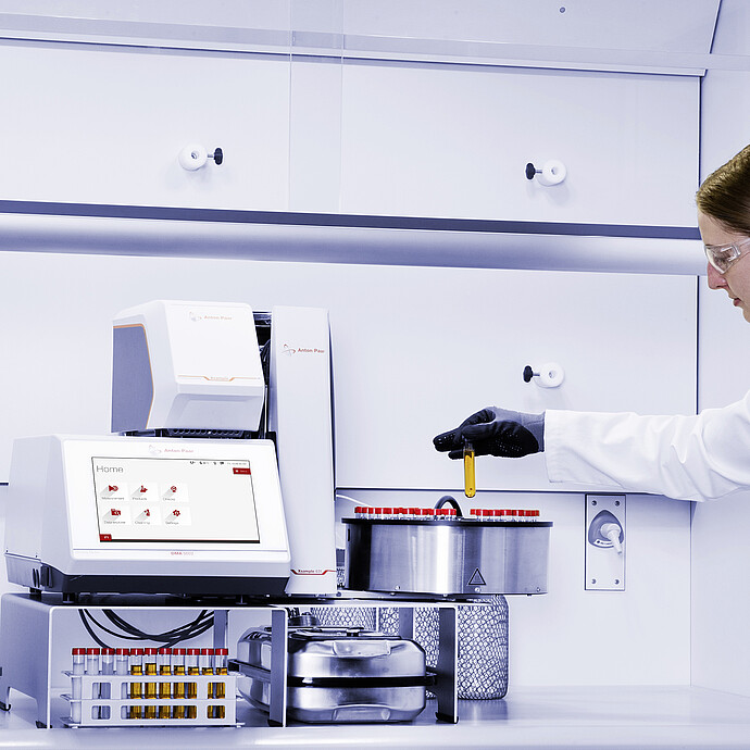 Lab technician holding a yellow vial beside Anton Paar DMA 5002 with Xsample Heating Controller and Xsample 631 sample changer carousel