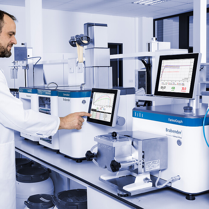 Technician operating Brabender ExtensoGraph and ExtensoBase, pointing at touchscreen with measurement curves, alongside Brabender FarinoGraph and Aquinject in laboratory