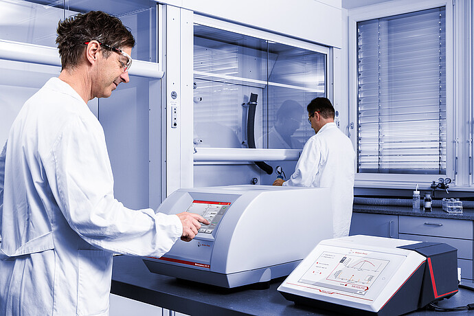 A scientist operates the Anton Paar Monowave 400 R microwave synthesis reactor via its touchscreen, while another scientist works in a laboratory fume hood. The Cora 5001 Raman spectrometer is positioned nearby, displaying molecular spectroscopy data on its screen.