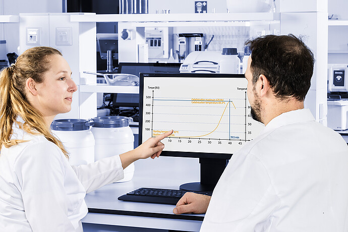 Two technicians reviewing viscosity curve on monitor, discussing measurement results from Brabender Amylograph-E in laboratory environment