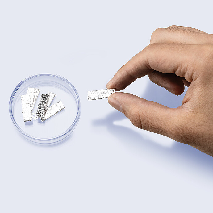 Hand holding a rectangular strip of eGaIn foil above a petri dish containing additional foil pieces for use in mercury-free porosimetry analysis.