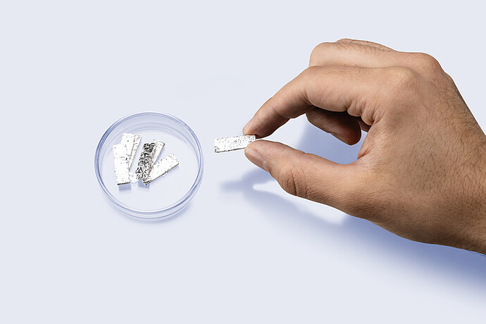 Hand holding a rectangular strip of eGaIn foil above a petri dish containing additional foil pieces for use in mercury-free porosimetry analysis.