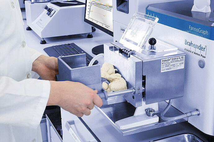 Close-up of technician loading dough into Brabender FarinoGraph mixing chamber, with open lid and visible kneading blades in laboratory setup