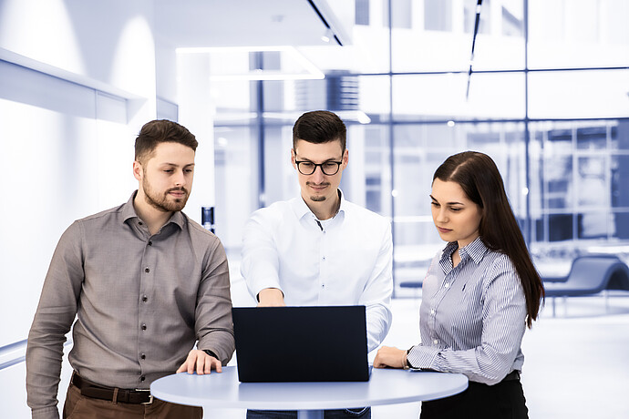 Three colleagues standing around a laptop in a modern office, collaborating during a business discussion.