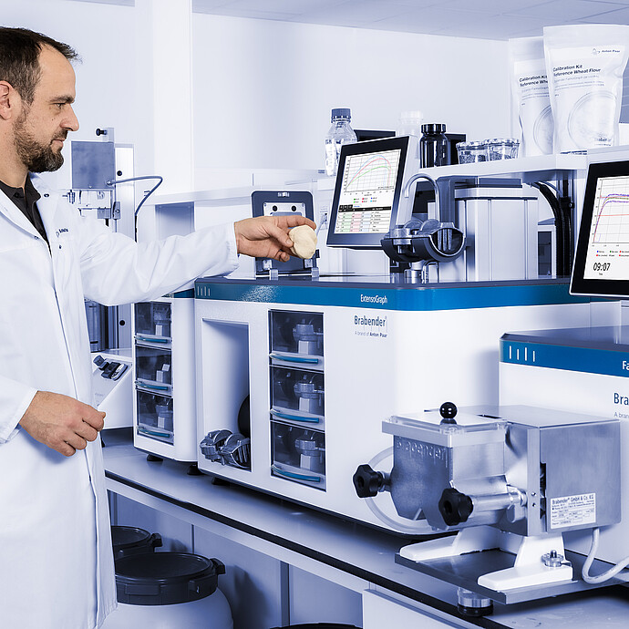 Technician placing dough sample into Brabender ExtensoGraph beside Brabender FarinoGraph, with monitors displaying measurement curves in a laboratory setting