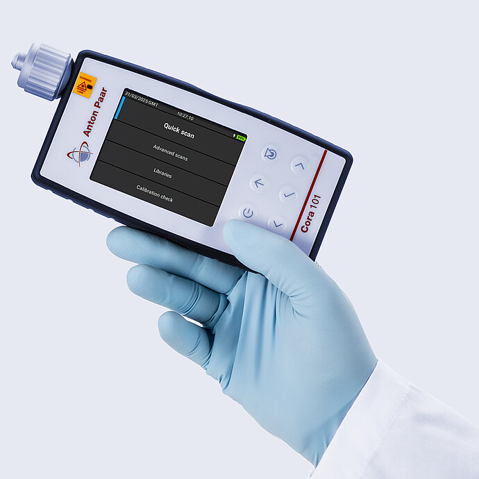 Gloved hand holding the Anton Paar Cora 101 handheld Raman spectrometer showing the main menu on its display screen.