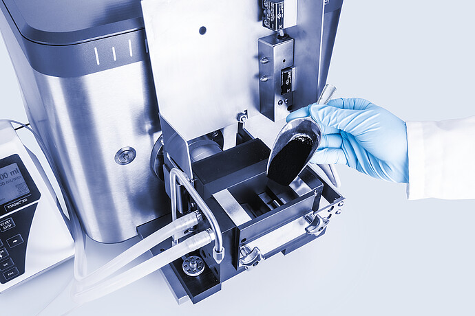 Close-up of Brabender AbsorptoMeter measuring chamber as gloved technician adds powder sample with scoop, sigma blades and tubing visible