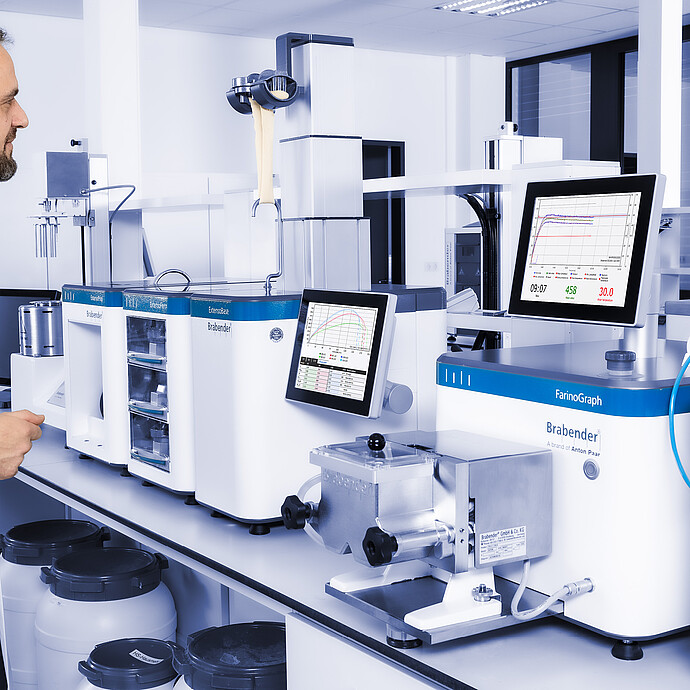 Technician observing dough stretching on Brabender ExtensoGraph with ExtensoBase, alongside Brabender FarinoGraph and Aquinject, monitors showing measurement curves in lab