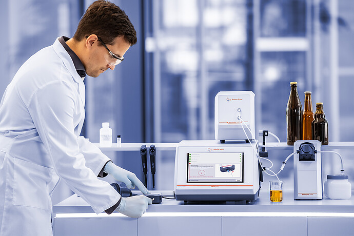 Lab technician using Anton Paar Enzymatic Reader with DMA 5002, Alcolyzer 3001, and Xsample 3100 beside beer bottles and sample beaker.