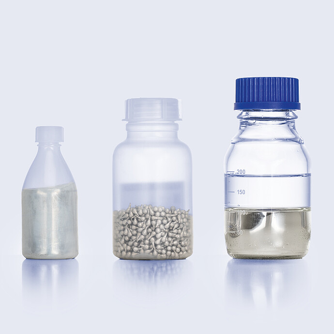 Three bottles with eGaIn shown in different forms: foil, pellets, and liquid metal in a lab bottle with volume markings.