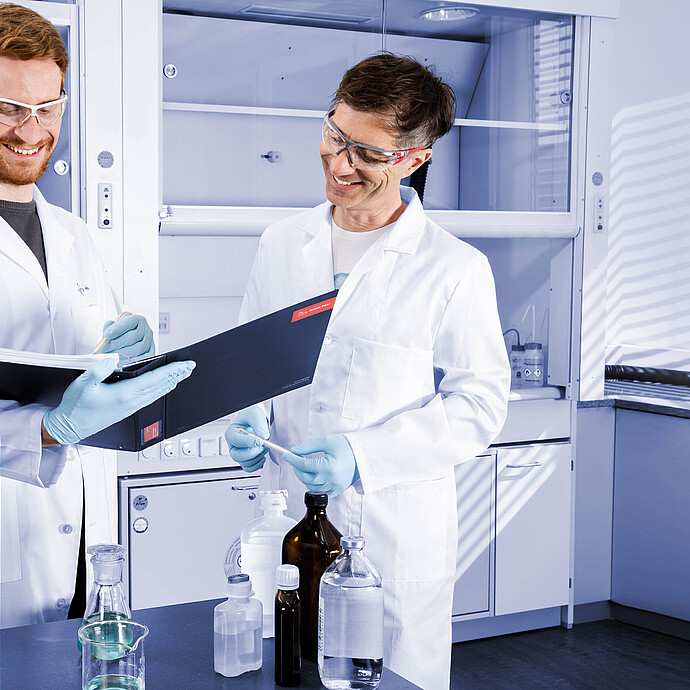 Two lab technicians in a laboratory reviewing Anton Paar-branded folders, with reagent bottles and lab glassware on the bench.