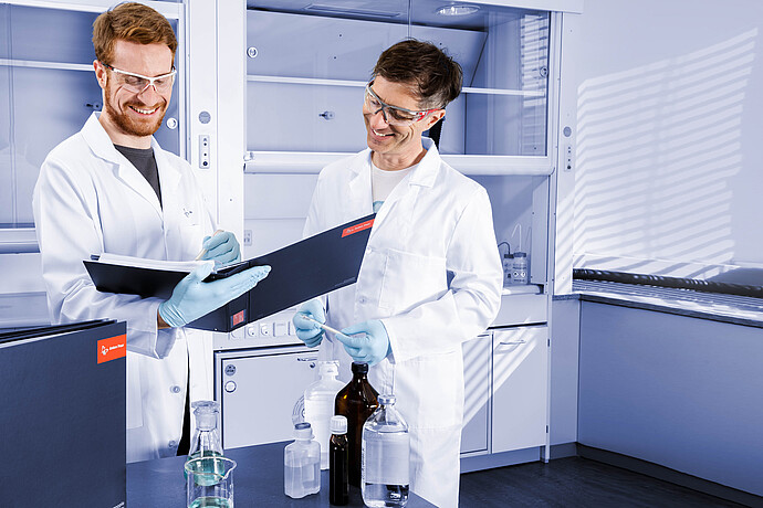 Two lab technicians in a laboratory reviewing Anton Paar-branded folders, with reagent bottles and lab glassware on the bench.