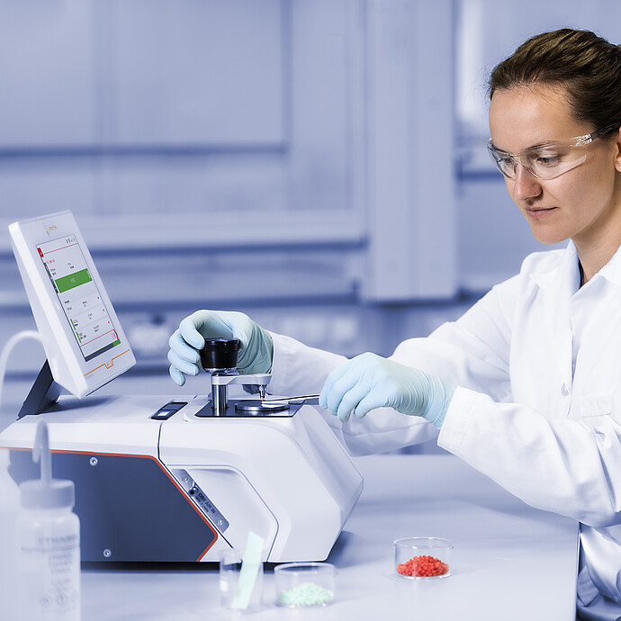Technician in safety glasses positions a sample with tweezers on an Anton Paar Lyza FTIR spectrometer beside red and green pellets.