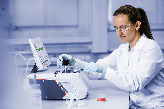 Technician in safety glasses positions a sample with tweezers on an Anton Paar Lyza FTIR spectrometer beside red and green pellets.