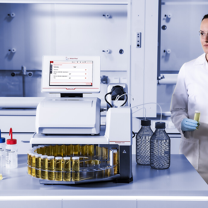 Laboratory technician holding sample tubes beside Anton Paar Lyza 7000 FTIR spectrometer with Xsample 5200 autosampler.