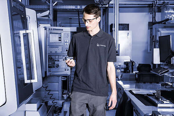 Anton Paar employee wearing safety glasses inspecting a component in a high-tech production facility with CNC machines and monitoring systems.