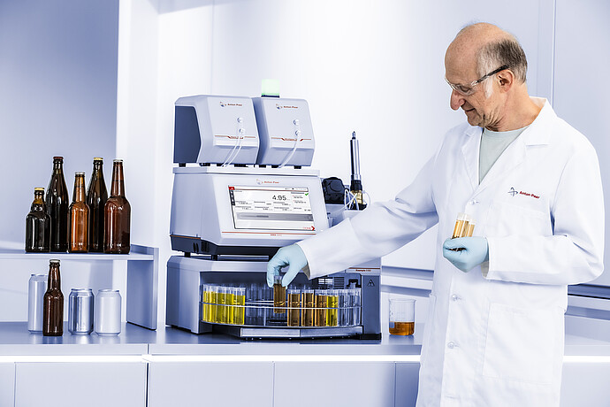 Technician handling beer samples beside Anton Paar RTD Measurement System with DMA 5002, HazeQC ME, Alcolyzer ME, and Xsample 5100.