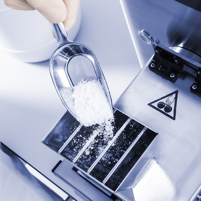 Close-up of Brabender AbsorptoMeter hopper as gloved hand adds white powder sample with scoop, metal inlet and safety symbol visible