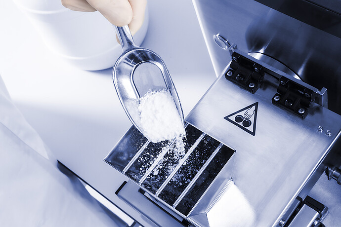 Close-up of Brabender AbsorptoMeter hopper as gloved hand adds white powder sample with scoop, metal inlet and safety symbol visible