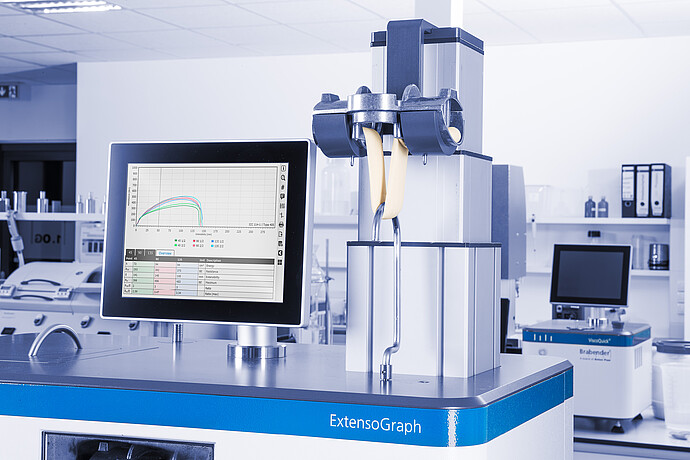 Brabender ExtensoGraph measuring stretched dough sample with clamps, touchscreen displaying extensibility curves, laboratory background with analytical instruments