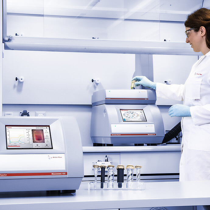 A scientist in a laboratory places a reaction vial into an Anton Paar Monowave 400 microwave synthesis reactor. Another Monowave 400 is on the lab bench, displaying reaction monitoring data. Several sealed reaction vials are arranged in the foreground.