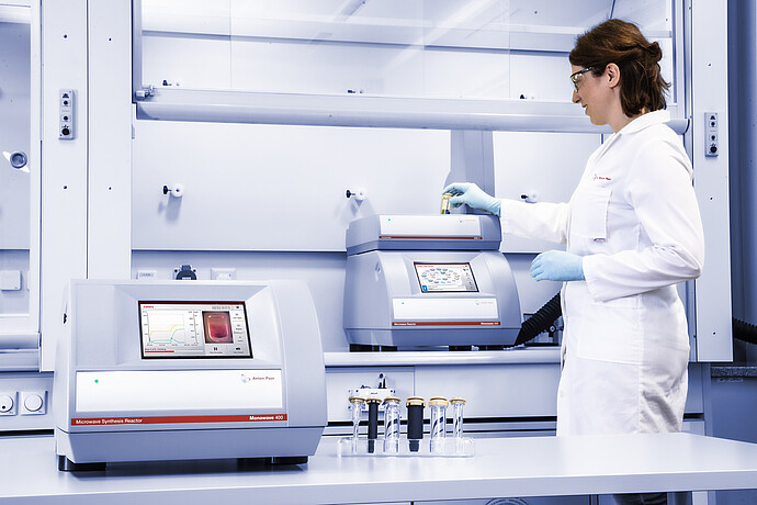 A scientist in a laboratory places a reaction vial into an Anton Paar Monowave 400 microwave synthesis reactor. Another Monowave 400 is on the lab bench, displaying reaction monitoring data. Several sealed reaction vials are arranged in the foreground.