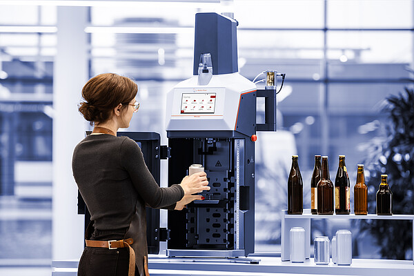 Technician places a beverage can into the Anton Paar TPO 5001 for total package oxygen measurement; bottles and cans are arranged on the side table.