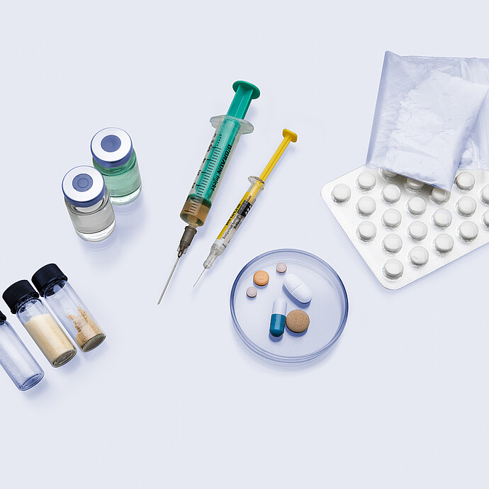 Various pharmaceutical samples including tablets, capsules, syringes, powder bags, vials with liquids and solids, and a petri dish on a white surface.