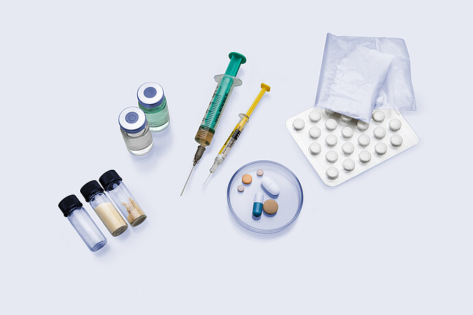 Various pharmaceutical samples including tablets, capsules, syringes, powder bags, vials with liquids and solids, and a petri dish on a white surface.