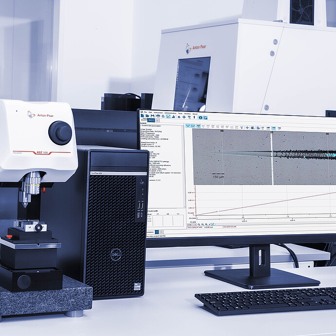 Anton Paar Revetest® Scratch Tester RST 100 setup on a laboratory desk, connected to a computer displaying scratch test results and analysis software on the monitor.