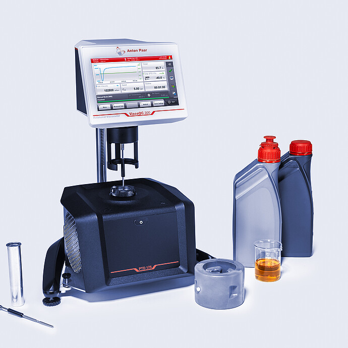Three-quarter front view of the ViscoQC 300 rotational viscometer with the PTD 175 Peltier temperature device, shown with oil bottles, a beaker of amber liquid, and accessories on a light blue background