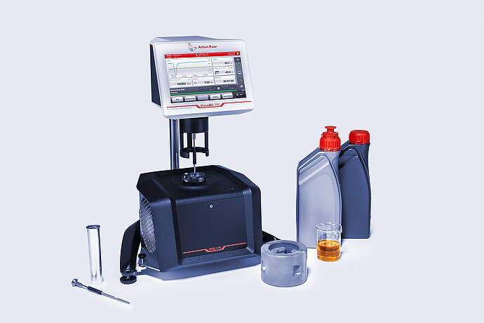 Three-quarter front view of the ViscoQC 300 rotational viscometer with the PTD 175 Peltier temperature device, shown with oil bottles, a beaker of amber liquid, and accessories on a light blue background