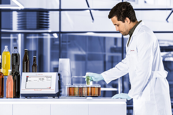Technician in lab coat operating an Anton Paar Alcolyzer 7001 Beer with beer samples in test tubes and various beverage bottles.